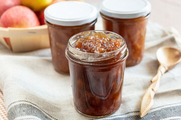 Homemade apple jam marmalade with cloves and cinnamon from organic apples in a transparent glass jar next to a carved wooden spoon. Homemade autumn jam preserves.