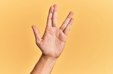 Arm and hand of caucasian man over yellow isolated background greeting doing vulcan salute, showing hand palm and fingers, freak culture