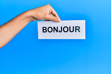 Hand of hispanic man holding french greeting bonjour word paper over isolated blue background. © Krakenimages.com