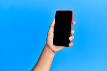 Hand of young hispanic man showing smartphone over isolated blue background.
