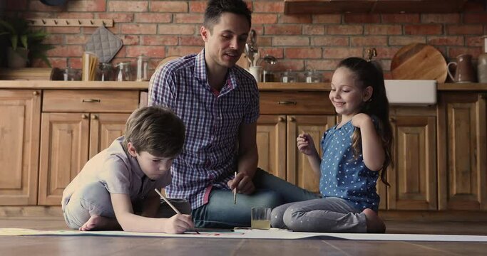 Full length joyful funny small girl and boy having fun, drawing with paintbrushes on paper sheet, enjoying spending free weekend leisure hobby activity time with loving young father in kitchen.