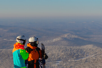 Two people in ski suits, masks and helmets stand on the top of a mountain on a sunny day. One of them is holding a snowboard