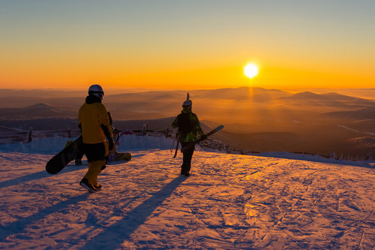 Sunset. The top of a snowy mountain. A snowboarder and a skier go down against the golden sunset