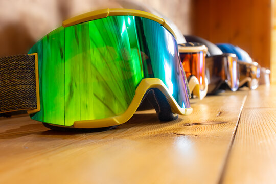 Ski goggles with different lenses are placed in a row one after the other. The first mask has a green lens, it is in focus. The ski goggles lie on a wooden surface