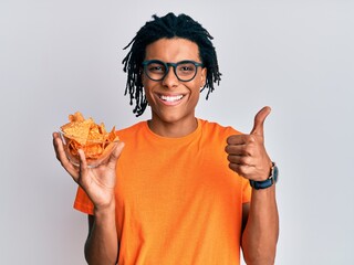 Young african american man holding nachos potato chips smiling happy and positive, thumb up doing...