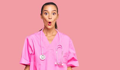 Young hispanic woman wearing doctor uniform and stethoscope scared and amazed with open mouth for surprise, disbelief face