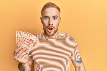 Young caucasian man holding mexican pesos scared and amazed with open mouth for surprise, disbelief face