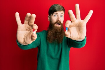 Redhead man with long beard holding two different pills clueless and confused expression. doubt concept.