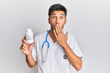 Young handsome man wearing doctor uniform holding presciption pills covering mouth with hand,...