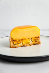 A yellow mango mousse tartelette cut in half on a white plate, a dessert fork, marble background. Light, bright, airy photo