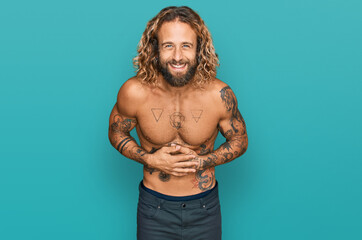 Handsome man with beard and long hair standing shirtless showing tattoos smiling and laughing hard out loud because funny crazy joke with hands on body.