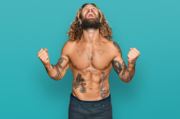 Obraz premium Handsome man with beard and long hair standing shirtless showing tattoos crazy and mad shouting and yelling with aggressive expression and arms raised. frustration concept.