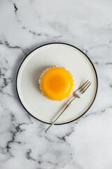 A yellow mango mousse tartelette on a white plate, a dessert fork, marble background. Light, bright, airy photo, top view