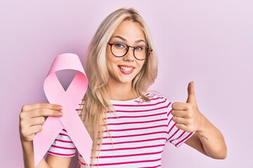 Beautiful caucasian blonde girl holding pink cancer ribbon smiling happy and positive, thumb up doing excellent and approval sign