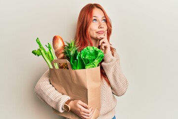 Young redhead woman holding paper bag with bread and groceries serious face thinking about question with hand on chin, thoughtful about confusing idea