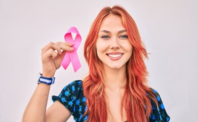 Young redhead girl smiling happy holding breast cancer pink ribbon walking at the city.
