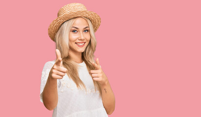 Young blonde girl wearing summer hat pointing fingers to camera with happy and funny face. good energy and vibes.