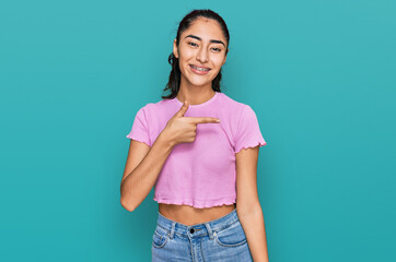 Hispanic teenager girl with dental braces wearing casual clothes cheerful with a smile on face pointing with hand and finger up to the side with happy and natural expression