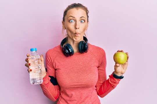 Beautiful caucasian sports woman holding water bottle and green apple making fish face with mouth and squinting eyes, crazy and comical.