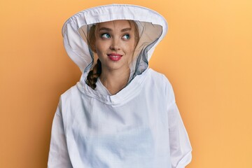 Beautiful blonde caucasian woman wearing protective beekeeper uniform smiling looking to the side and staring away thinking.