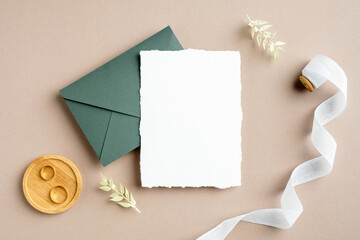 Wedding invitation card mockup, green envelope, golden rings, silk ribbon, dried flowers on pastel beige background. Flat lay, top view, copy space. Rustic wedding stationery set.