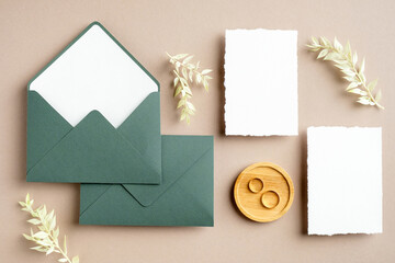Feminine wedding stationery set. Blank greeting card, green envelopes, dried flowers, golden rings on pastel beige background. Rustic wedding invitation cards. Flat lay, top view, copy space.