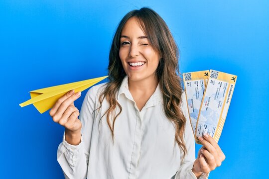 Young brunette woman holding paper airplane and boarding pass winking looking at the camera with sexy expression, cheerful and happy face.