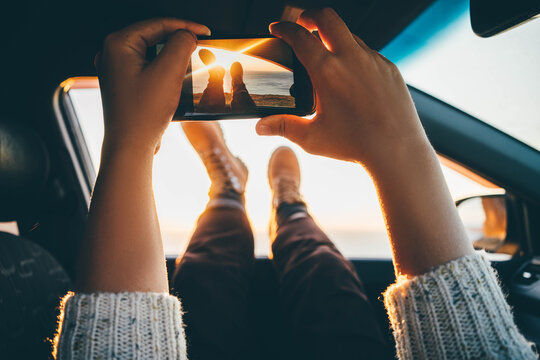 Woman taking pictures with smartphone out of car window at sunset. Travel concept.