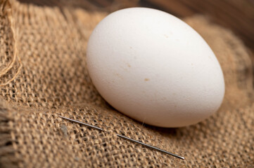 A chicken egg and a sewing needle on a homespun fabric with a rough texture.