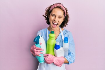 Young brunette woman wearing cleaner apron holding cleaning products smiling and laughing hard out...