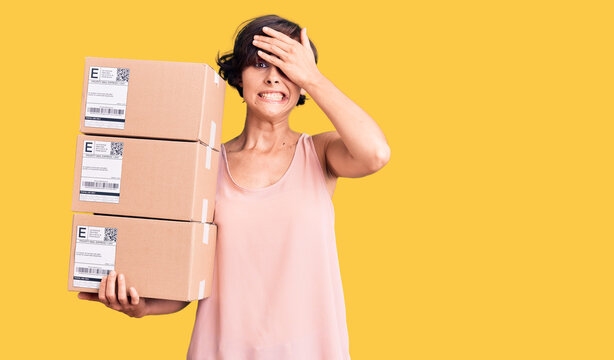 Beautiful young woman with short hair holding delivery packages stressed and frustrated with hand on head, surprised and angry face