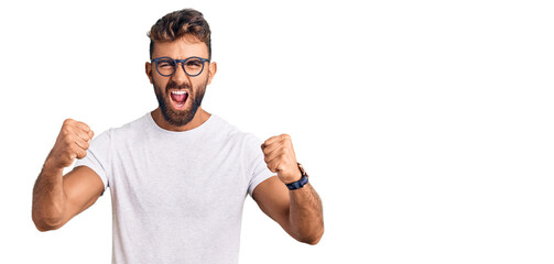 Young hispanic man wearing casual clothes and glasses angry and mad raising fists frustrated and...