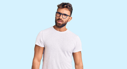 Young hispanic man wearing casual clothes and glasses in shock face, looking skeptical and...