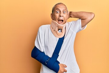 Handsome mature senior man wearing cervical collar and arm on sling crazy and scared with hands on...