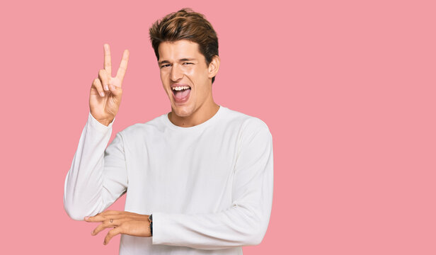 Handsome caucasian man wearing casual white sweater smiling with happy face winking at the camera doing victory sign with fingers. number two.