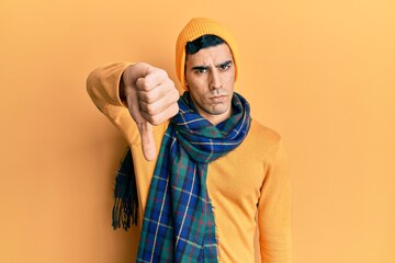 Handsome hispanic man wearing wool hat and winter scarf looking unhappy and angry showing rejection and negative with thumbs down gesture. bad expression.