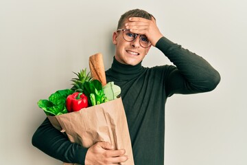 Young caucasian man holding paper bag with groceries stressed and frustrated with hand on head, surprised and angry face