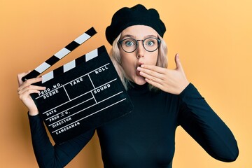 Young blonde girl holding video film clapboard covering mouth with hand, shocked and afraid for...
