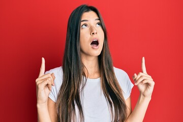 Young brunette woman wearing casual white tshirt over red background amazed and surprised looking up and pointing with fingers and raised arms.