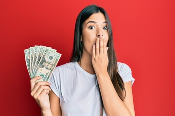 Young brunette woman holding 20 dollars banknote covering mouth with hand, shocked and afraid for mistake. surprised expression