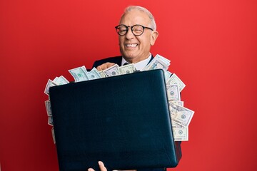 Senior caucasian man holding briefcase full of dollars winking looking at the camera with sexy...