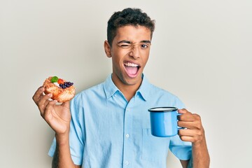 Young handsome african american man drinking coffee and eating pastry winking looking at the camera...
