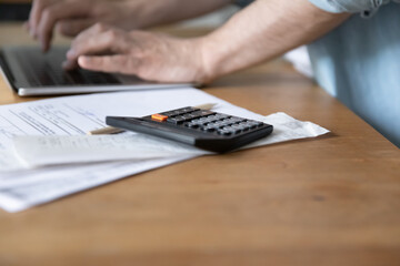 Close up of male client user of electronic bank system typing on laptop keyboard making payment online. Focus on calculator device lying on financial paper documents stack bills invoices notifications