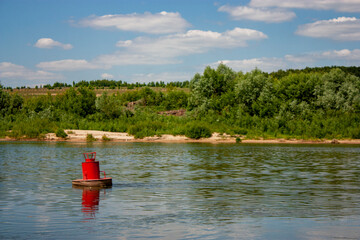 View from the river bank to the red buoy on a bright summer day. The red buoy is reflected in the calm water of the river. Blue sky and clouds are reflected in the river.
