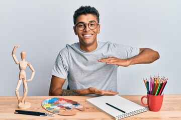 Young handsome african american man painter sitting palette and art manikin gesturing with hands showing big and large size sign, measure symbol. smiling looking at the camera. measuring concept.
