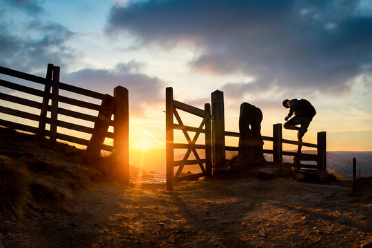 Silhouette of anonymous hiker lone man climbing fence at beautiful orange sunrise on horizon in Peak District with open gate casting shadow on Mam Tor ridge Derbyshire