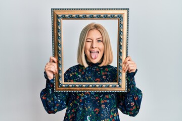 Fototapeta premium Young blonde woman holding empty frame sticking tongue out happy with funny expression.