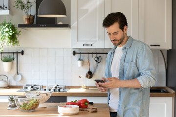 Millennial male distracted from preparing food hold smartphone order missing ingredients online. Young man husband stand at modern kitchen using cell consulting with wife about cooking process details