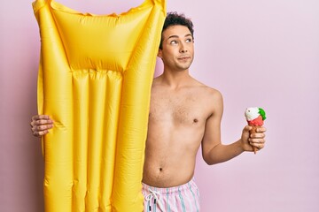 Young handsome man wearing swimsuit holding summer mattress float and icecream smiling looking to the side and staring away thinking.