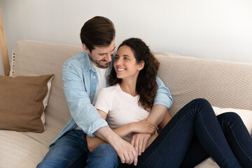 Dreamful young family couple hug on cozy sofa relaxing at living room on weekend chatting discussing future plans. Happy young husband sit on couch hold dear wife in arms having pleasure in speaking